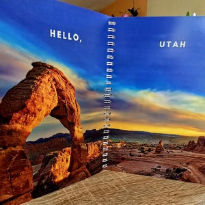 May include: An open notebook with a blue cover and a spiral binding. The left page features a photo of a large sandstone arch with the text "HELLO," and the right page features a photo of a desert landscape with the text "UTAH."
