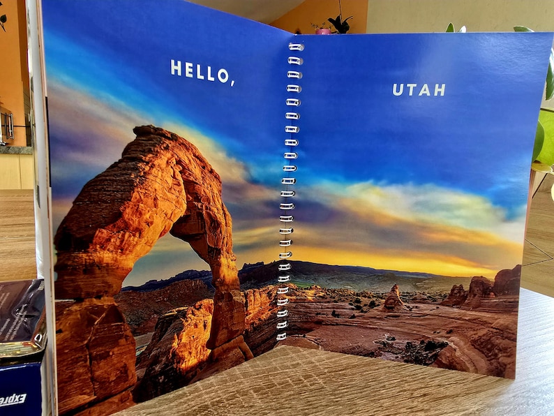 May include: An open notebook with a blue cover and a spiral binding. The left page features a photo of a large sandstone arch with the text "HELLO," and the right page features a photo of a desert landscape with the text "UTAH."