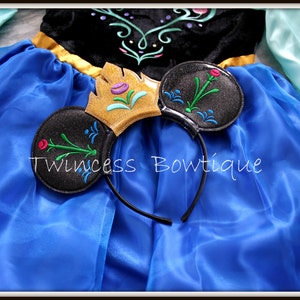 May include: A blue dress with long light blue sleeves and a black bodice featuring floral embroidery, paired with a black headband featuring two round ears, a gold crown, and floral embroidery. The text Twincess Bowtique is visible.