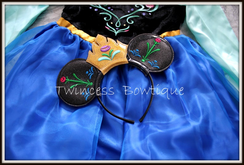May include: A blue dress with long light blue sleeves and a black bodice featuring floral embroidery, paired with a black headband featuring two round ears, a gold crown, and floral embroidery. The text Twincess Bowtique is visible.