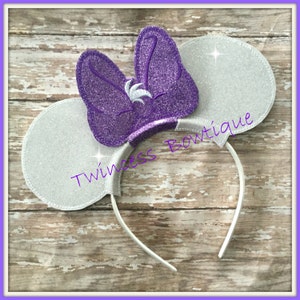 May include: Silver glitter mouse ears with a purple glitter bow. The bow has a white swirl design. The headband is white.