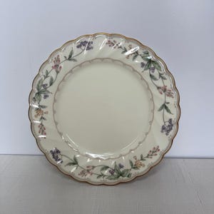 Peut inclure: Une assiette ronde en céramique blanc cassé avec un bord festonné et un bord doré. L'assiette est ornée d'un motif floral dans les tons de rose, de violet et de vert. Le design est délicat et élégant, adapté aux dîners formels.