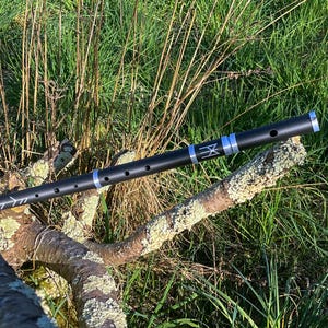 Peut inclure: Une flûte en bois noir avec des accents argentés et un symbole de rune sur le corps. La flûte repose sur une branche recouverte de mousse verte.