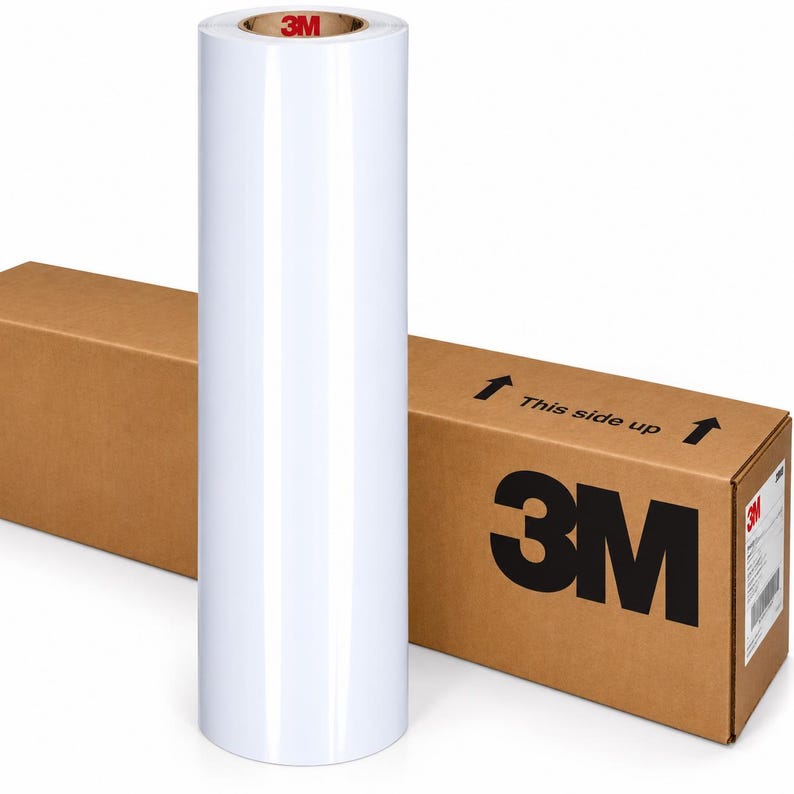 May include: A tall, cylindrical roll of glossy white adhesive film standing upright. The core of the roll features the red 3M logo. Behind the roll is a rectangular brown cardboard shipping box with the 3M logo and the text This side up printed.