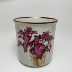 May include: A white ceramic mug with a brown rim and a floral design. The design features a potted plant with pink flowers and green leaves.