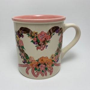 May include: Cream-colored ceramic mug with a pink interior and handle. The mug features a heart-shaped floral design with pink, orange, and purple flowers. The design is accented with brown ribbons and pink decorative elements.