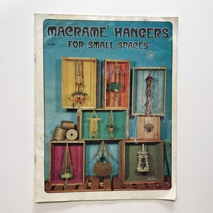 May include: A vintage book cover featuring the title "Macrame' Hangers for Small Spaces" and a colourful illustration of macrame plant hangers displayed in wooden crates.