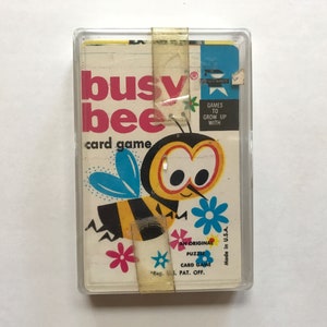 以下が含まれることがあります： 黄色と黒の漫画の蜂のイラストが描かれたビンテージのカードゲームの箱。箱には「busy bee card game」と表示され、「Games to grow up with」というテキストが書かれています。
