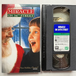 May include: VHS tape case with a black background and a blue label that reads "Miracle on 34th Street". The front cover of the case features a photo of Santa Claus and a young girl.