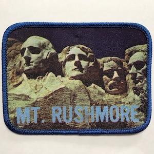 May include: A blue and white embroidered patch featuring a photo of Mount Rushmore National Memorial. The patch shows the four presidents carved into the mountain: George Washington, Thomas Jefferson, Theodore Roosevelt, and Abraham Lincoln. The text "MT. RUSHMORE" is printed in blue below the image.