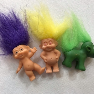 May include: Three small troll dolls with colourful hair. One troll has purple hair, one has yellow hair, and one has green hair. The troll with green hair is riding a green troll.