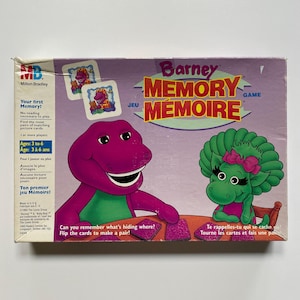 May include: A Barney Memory Game box featuring Barney the dinosaur and a green dinosaur character. The box has the text "Memory Game" and "Jeu Mémoire". The game is for ages 3 to 6 and requires no reading to play.