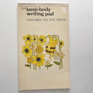 May include: A yellow and black bee with a bucket hanging from its leg is surrounded by yellow flowers with black centers. The text "The busy-body writing pad" and "Stationery for busy people" is printed on the front of the pad.