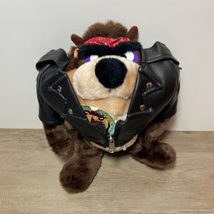 May include: A plush Tasmanian Devil toy wearing a black faux leather jacket with silver-tone metal snaps and a red patterned bandana. The character has brown fur, a large black nose, and purple eyelids. A circular patch featuring the character is visible.