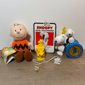 May include: A collection of Snoopy and Peanuts-themed toys and collectibles. Includes a Charlie Brown plush doll, a Snoopy telephone, a yellow Woodstock figurine, and a Snoopy figure on a blue and yellow drum.