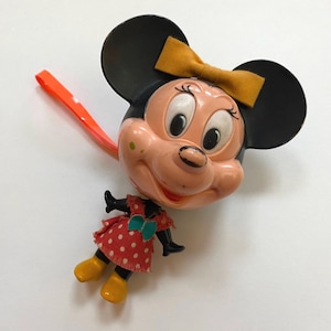 May include: A vintage plastic Minnie Mouse toy with a red and white polka dot dress, black shoes, and a yellow bow. The toy has a pull string and is in good condition.