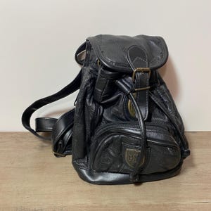 May include: Black leather backpack with a flap closure and adjustable straps. The small backpack features a front zippered pocket and a decorative emblem. The bag has a vintage aesthetic.