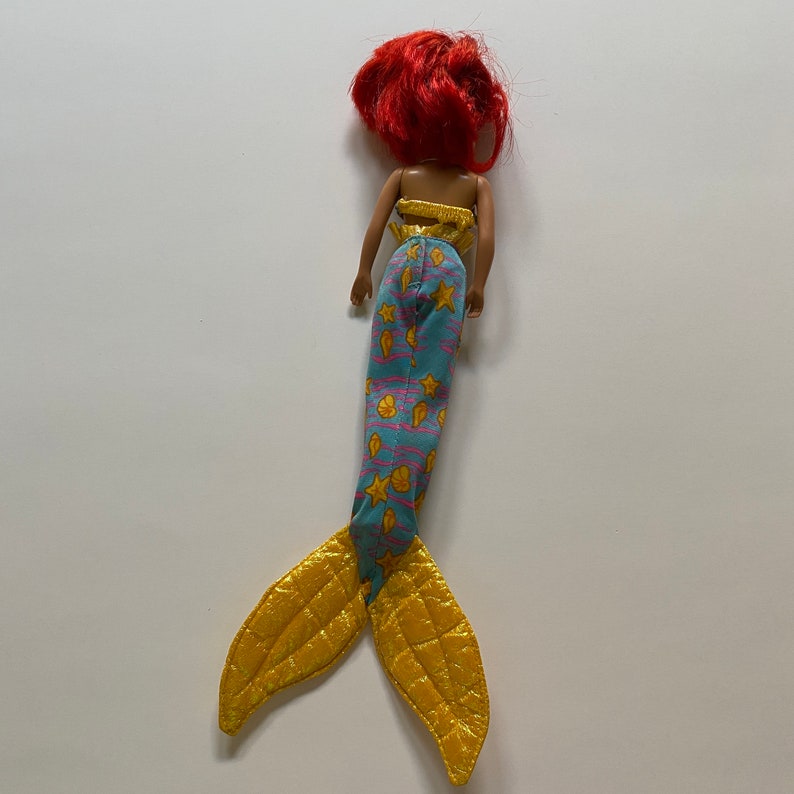 Puede incluir: Una mu&ntilde;eca sirena con cabello rojo, una cola azul y rosa con conchas y estrellas amarillas, y una aleta amarilla.
