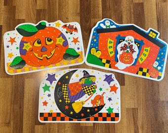 Puede incluir: Tres manteles individuales rectangulares con temática de Halloween y bordes redondeados. Uno presenta una gran calabaza naranja, otro muestra un fantasma blanco sosteniendo una calabaza en una casa y el tercero representa una bruja.