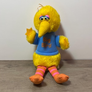 May include: A yellow Big Bird plush toy wearing a blue shirt with the words "Big Bird" in orange letters. The toy has pink and orange striped legs and is sitting on a wooden surface.