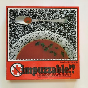 May include: A red jigsaw puzzle box with a soup bowl and spoon design. The puzzle is titled "Impuzzable!!" and features a 550-piece count. The box has a black and white speckled background and the text "too hot to handle".