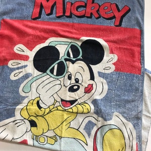 Pode incluir: Uma toalha de praia azul com borda vermelha e branca. O Mickey Mouse est&aacute; usando &oacute;culos de sol e uma camisa amarela. A toalha diz "Mickey" em letras vermelhas.
