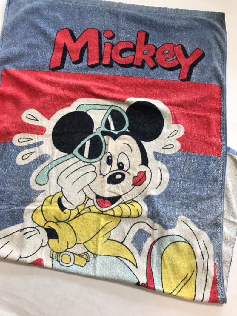 Pode incluir: Uma toalha de praia azul com borda vermelha e branca. O Mickey Mouse est&aacute; usando &oacute;culos de sol e uma camisa amarela. A toalha diz "Mickey" em letras vermelhas.