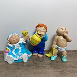May include: Three Cabbage Patch Kids figurines. A girl in a blue dress holds a rattle, a boy in blue overalls holds a yellow cookie jar, and a baby in a nappy holds a yellow towel.