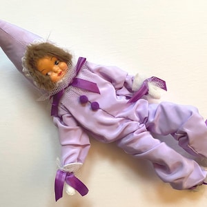 Peut inclure: Une poupée vêtue d'un costume de jester violet et blanc avec un chapeau pointu et un col à volants. La poupée a les cheveux bruns et un visage peint.
