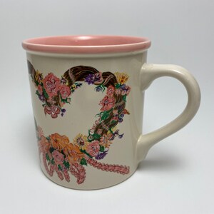 May include: Cream-colored ceramic mug with a pink interior rim and handle. The mug features a floral heart design with roses, sunflowers, and the name "Christine" printed on the side. A decorative coffee mug.