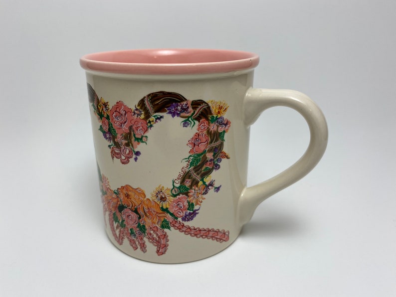 May include: Cream-colored ceramic mug with a pink interior rim and handle. The mug features a floral heart design with roses, sunflowers, and the name "Christine" printed on the side. A decorative coffee mug.