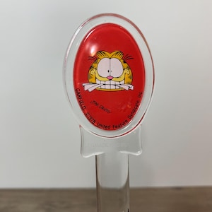 May include: A clear plastic Garfield character lollipop stick with a red oval top featuring Garfield's face. The text "GARFIELD ©1978 United Feature Syndicate, Inc." is printed around the oval.