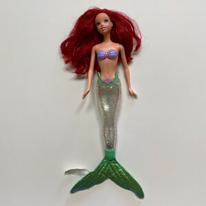 May include: A doll of a mermaid with red hair, blue eyes, and a purple seashell top. The doll has a clear, glitter-filled tail and a green, textured fin. The doll is a popular children's toy.