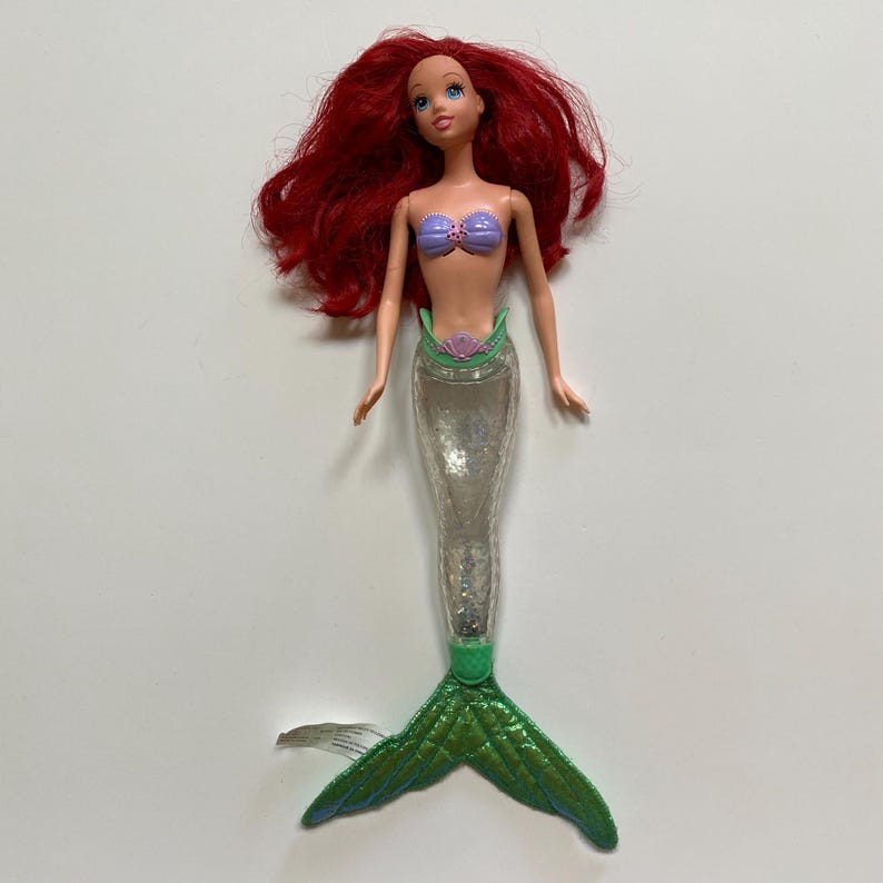 May include: A doll of a mermaid with red hair, blue eyes, and a purple seashell top. The doll has a clear, glitter-filled tail and a green, textured fin. The doll is a popular children's toy.