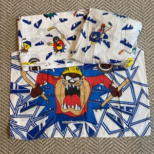 May include: A white bed sheet set featuring Looney Tunes characters playing hockey. The pillowcase displays the Tasmanian Devil holding broken hockey sticks against a blue geometric background. The sheets feature Tweety Bird and Yosemite Sam.