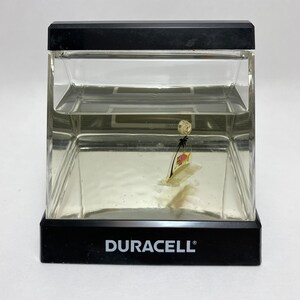 May include: A clear plastic cube with a black base and a small plastic sailboat inside. The sailboat has a palm tree and a red fish on it. The base of the cube has the word "DURACELL" printed on it.