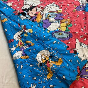 May include: A blue and pink quilted blanket with a Disney character theme. The blanket features Mickey Mouse, Donald Duck, Goofy, and Pluto. The characters are surrounded by white flower petals.