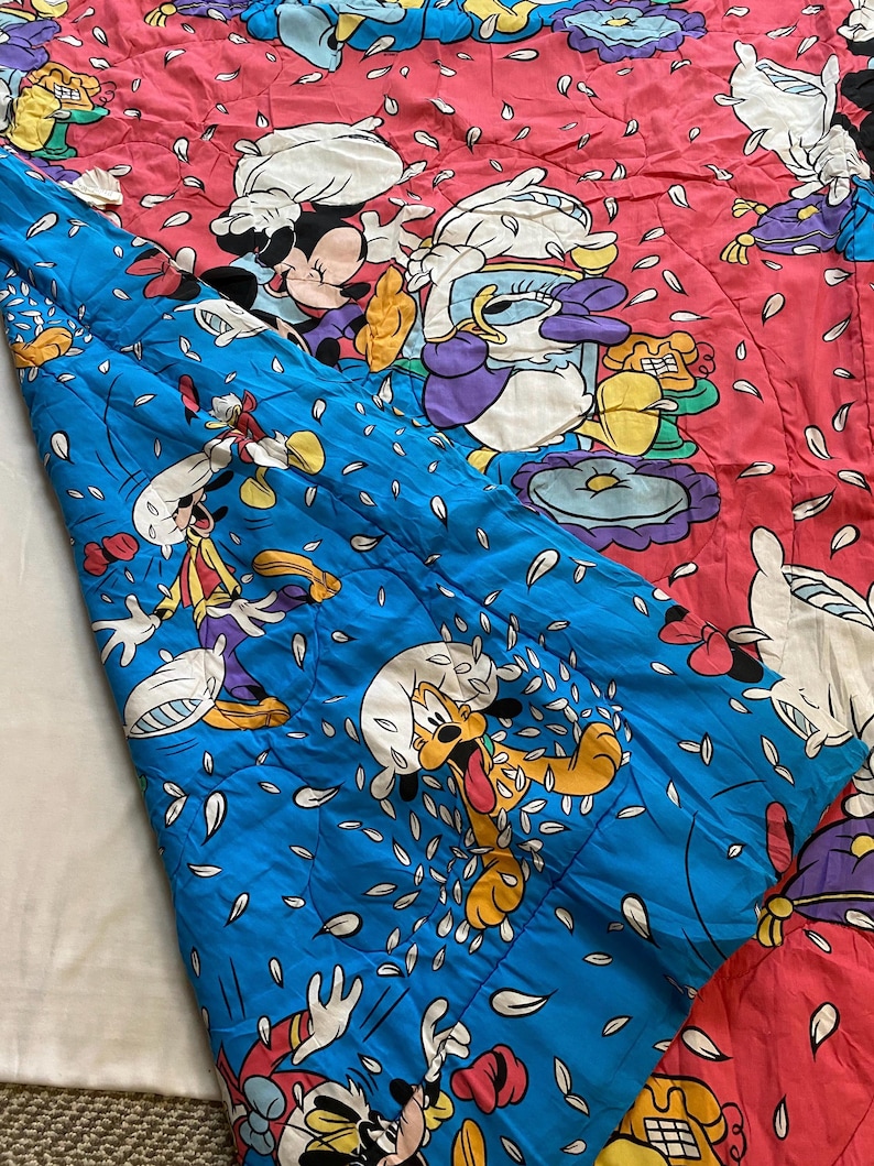 May include: A blue and pink quilted blanket with a Disney character theme. The blanket features Mickey Mouse, Donald Duck, Goofy, and Pluto. The characters are surrounded by white flower petals.