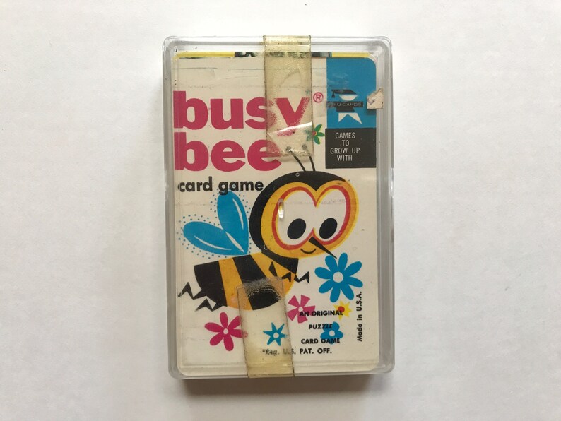 以下が含まれることがあります： 黄色と黒の漫画の蜂のイラストが描かれたビンテージのカードゲームの箱。箱には「busy bee card game」と表示され、「Games to grow up with」というテキストが書かれています。