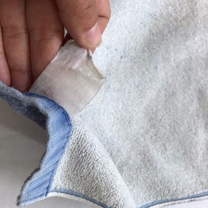 Pode incluir: Close-up de um moletom azul claro com uma etiqueta branca anexada. A etiqueta &eacute; parcialmente vis&iacute;vel e tem algum texto.