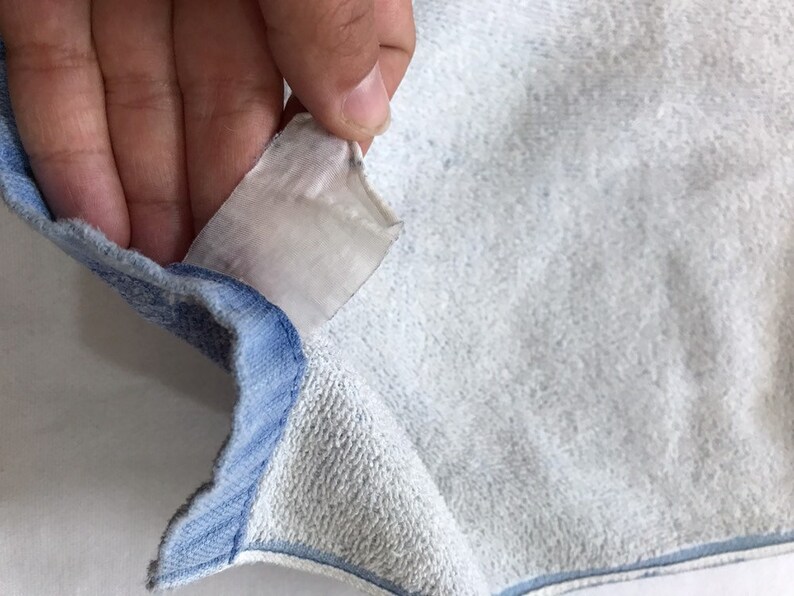 Pode incluir: Close-up de um moletom azul claro com uma etiqueta branca anexada. A etiqueta &eacute; parcialmente vis&iacute;vel e tem algum texto.