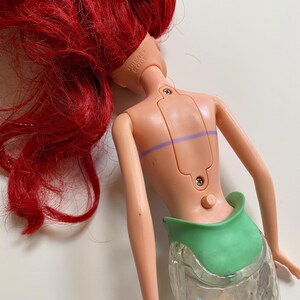 May include: A doll with red hair and a green tail. The doll's back is visible, showing a light pink plastic body with a green waist and a clear, textured tail. The doll's head is marked "Disney 2005".