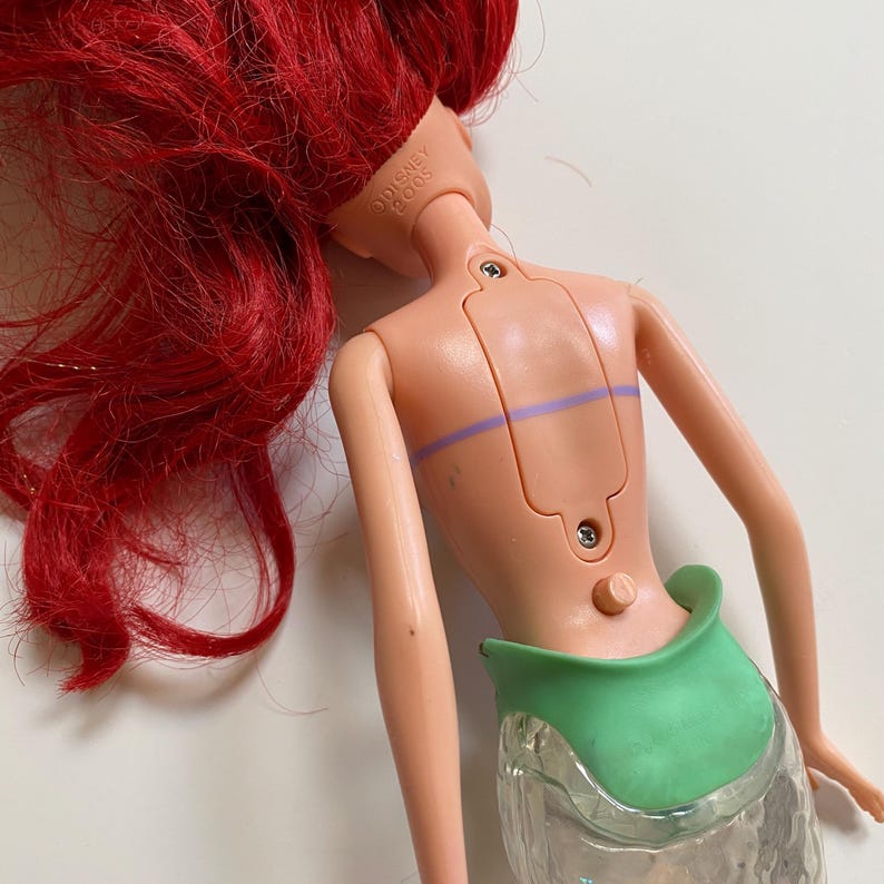 May include: A doll with red hair and a green tail. The doll's back is visible, showing a light pink plastic body with a green waist and a clear, textured tail. The doll's head is marked "Disney 2005".