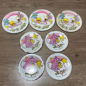 May include: A collection of seven round metal plates featuring vintage illustrations of a blonde child, a blue dog, and a purple cat on clouds with rainbows and yellow stars. Some plates show signs of wear and rust along the edges and surfaces.