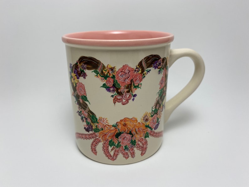 May include: Cream-colored ceramic mug with a pink interior and handle. The mug features a heart-shaped floral design with pink, orange, and purple flowers. The design is accented with brown ribbons and pink decorative elements.