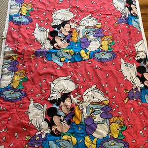 May include: A red and white quilt with a repeating pattern of Mickey Mouse and Donald Duck. The characters are surrounded by white flower petals.