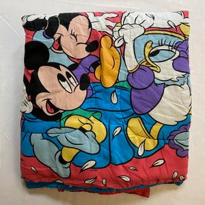 May include: A pink, blue, yellow, and white quilted blanket featuring Mickey Mouse and Minnie Mouse with a duck character. The blanket has a white background with pink flower petals scattered across it.