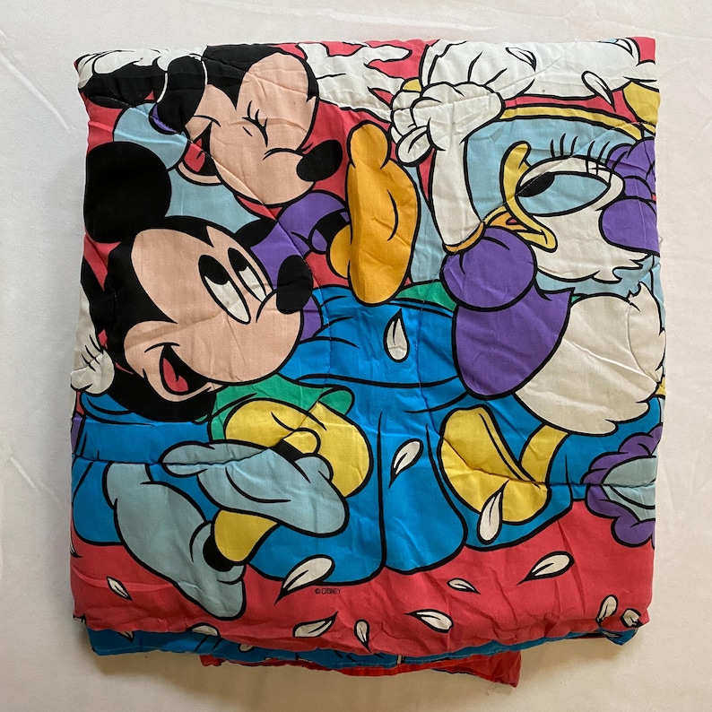 May include: A pink, blue, yellow, and white quilted blanket featuring Mickey Mouse and Minnie Mouse with a duck character. The blanket has a white background with pink flower petals scattered across it.