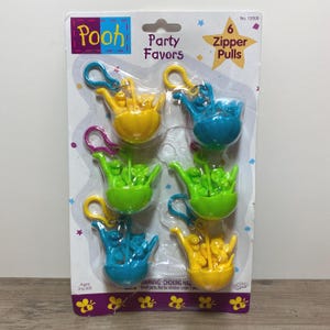 May include: A pack of six Winnie the Pooh zipper pulls. Each pull features a yellow, blue or green plastic cup with three Winnie the Pooh characters inside. The pack is labelled "Pooh Party Favors" and "6 Zipper Pulls".