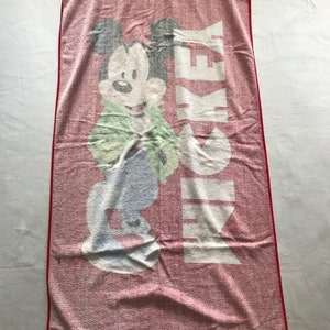 May include: A red and white Mickey Mouse beach towel. The towel features a large graphic of Mickey Mouse in a green jacket and blue pants. The word "MICKEY" is printed vertically in white. The towel has a red border.